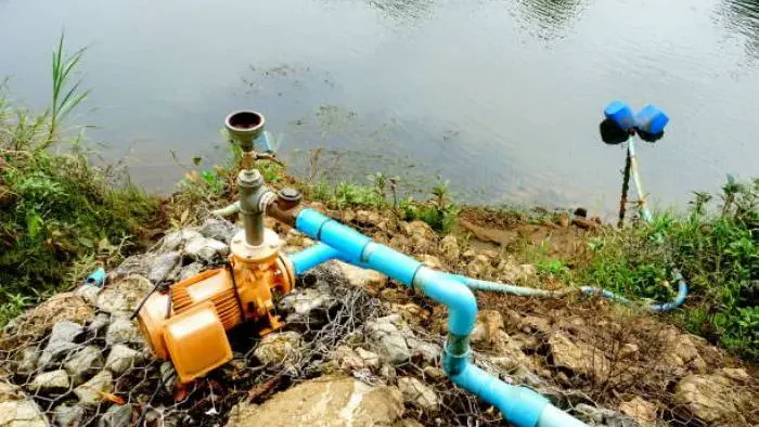 Water pump machinery connected to blue pipes, positioned near a pond, intended for water circulation.