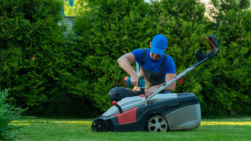 Lawn Mower Is Burning Oil - 5 Causes & How To Fix 2 Man performing maintenance on a lawn mower, possibly due to it burning oil, in a garden.