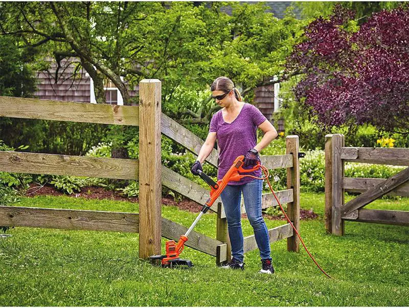 Woman operating an electric weed wacker along a wooden fence line.