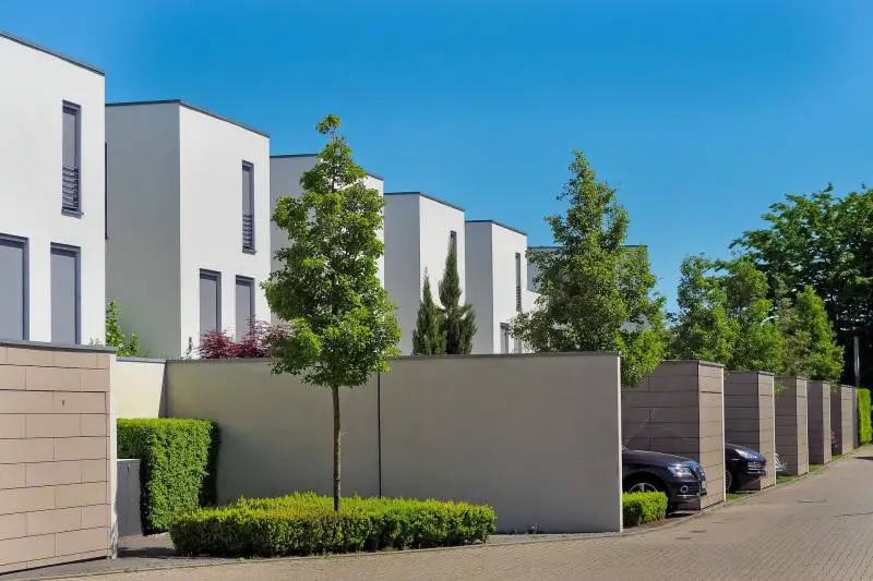 Modern residential street with uniform, cube-shaped houses and a tall neighbor's wall, lined with neatly trimmed hedges.