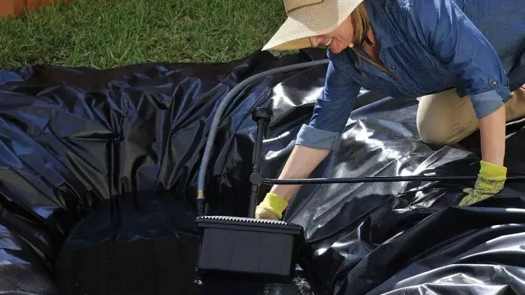 Woman in a garden hat installing a pond skimmer, spreading a black liner in a dug-out pond area to keep water clean and free of debris.